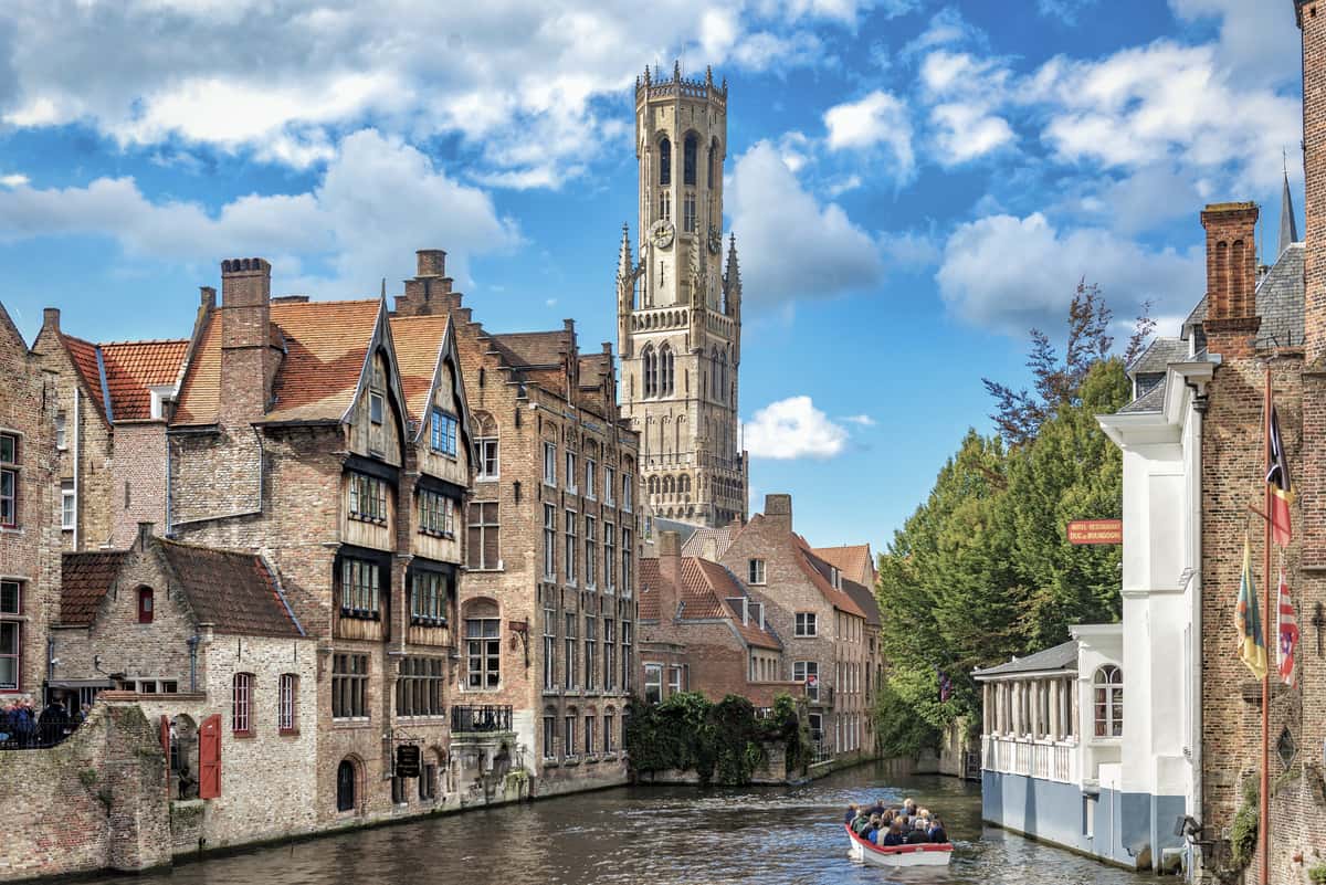 Cityscape with cathedral, a boat passing by on the river, and blue sky. Bruges and the Belfry