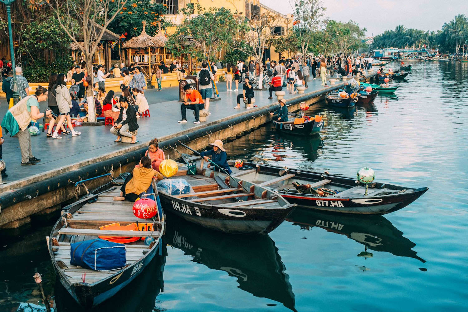 Old city of Hoi An with the canal, the tourists walking on the pavement, and 3 boats on the water during golden hour
