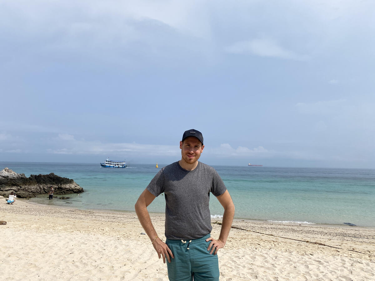 Man posing on a beach with a boat in the background.