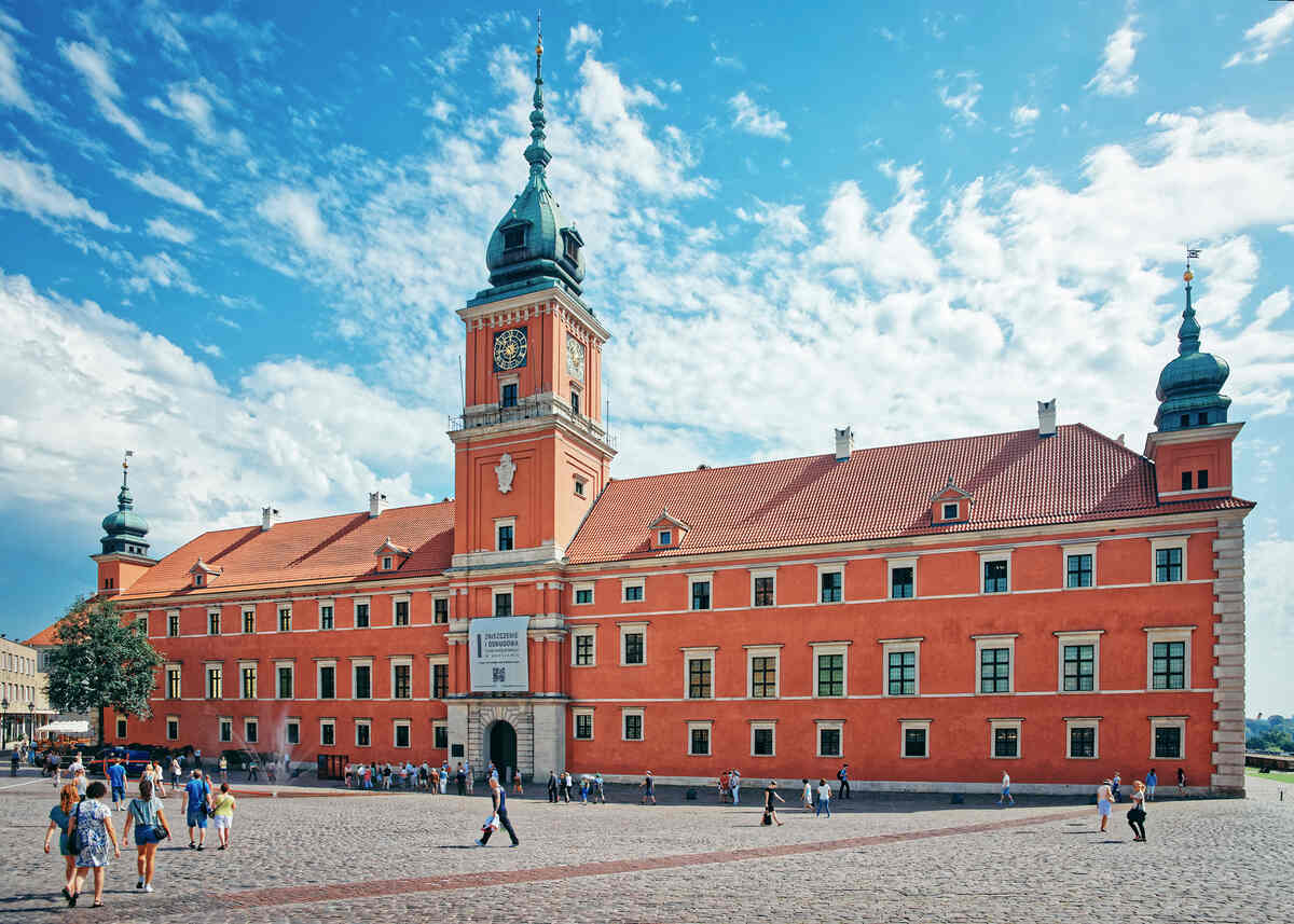 Enter the Royal Castle in Warsaw - A historic red-brick castle complex with a tower under a sunny sky.