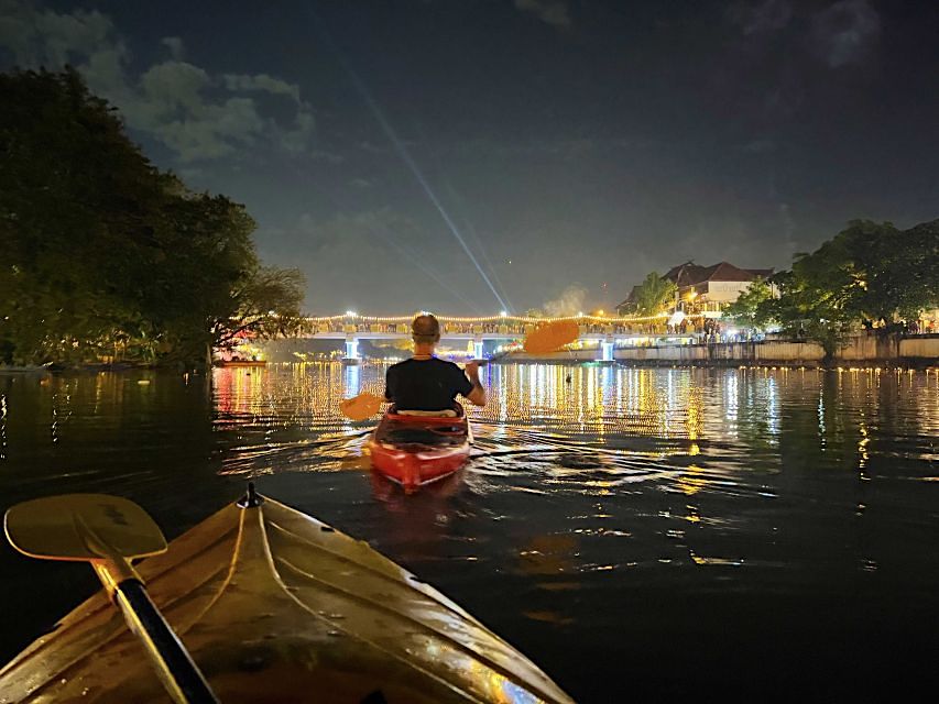 People in a kayak on a calm river at night, with city lights reflecting on the water.