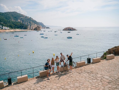 A panoramic view of a Mediterranean cove with boats in the azure water and tourists enjoying the sun on a coastal walkway.