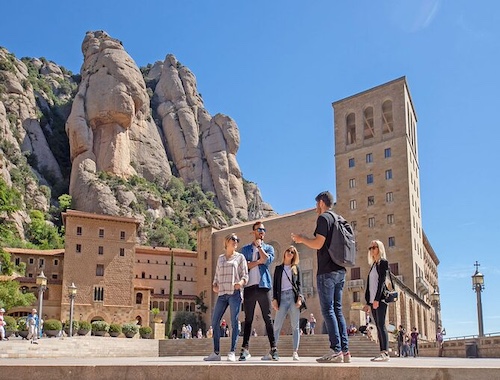 A group of tourists standing in front of a towering, rocky mountain formation with a historic building integrated into the landscape.