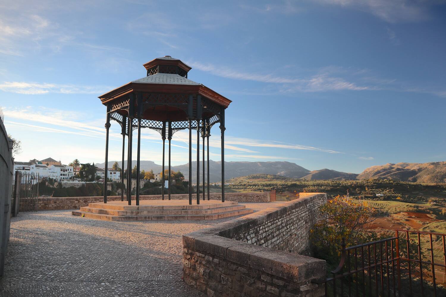 A gazebo on a clear day with a bench overlooking a panoramic view of a hilly landscape.
