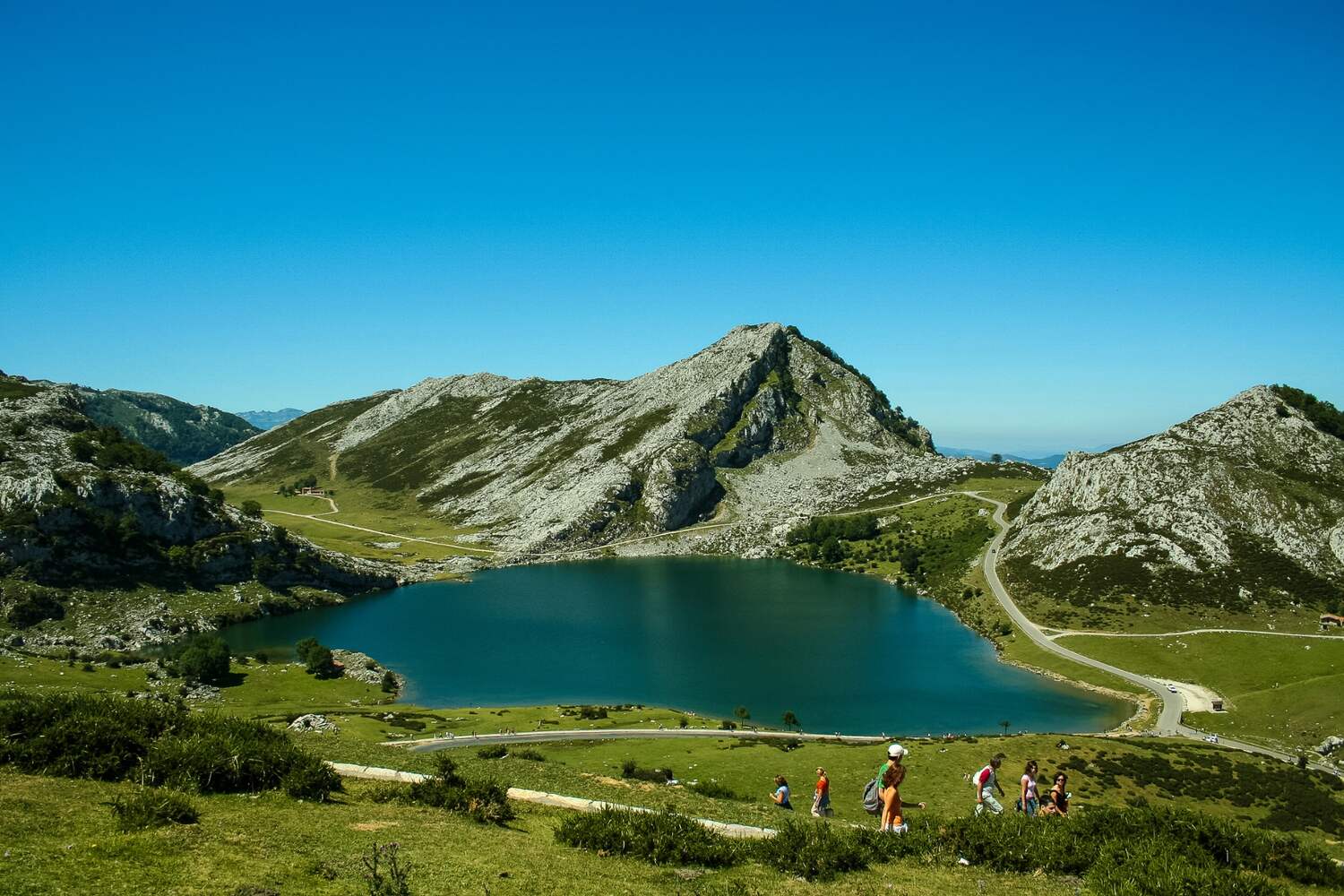 Enol Lake in the Picos de Europa National Park, Spain, surrounded by lush green slopes and a clear blue sky.
