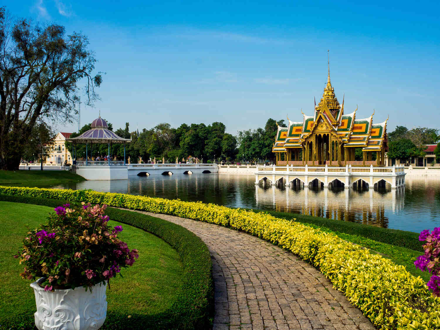 A scenic park with a large pond, vibrant flower beds, and a traditional Thai pavilion in the distance.