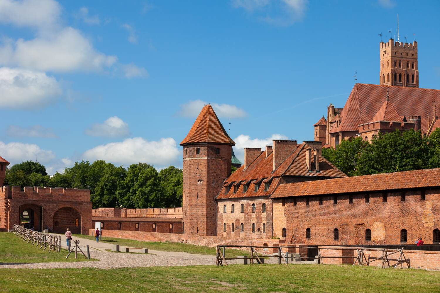 Discover-the-Malbork-Castle, A medieval brick castle with defensive towers, surrounded by a dry moat and green lawns under a clear blue sky.
