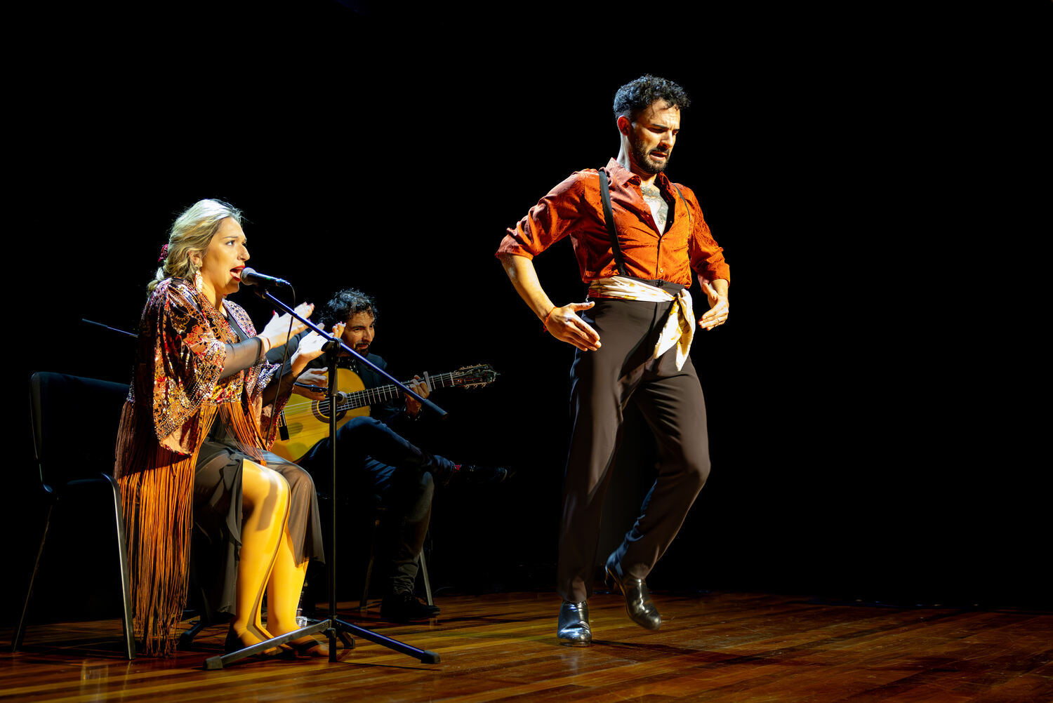 Man dancing Flamenco in a black room with a man playing guitar and a woman singing