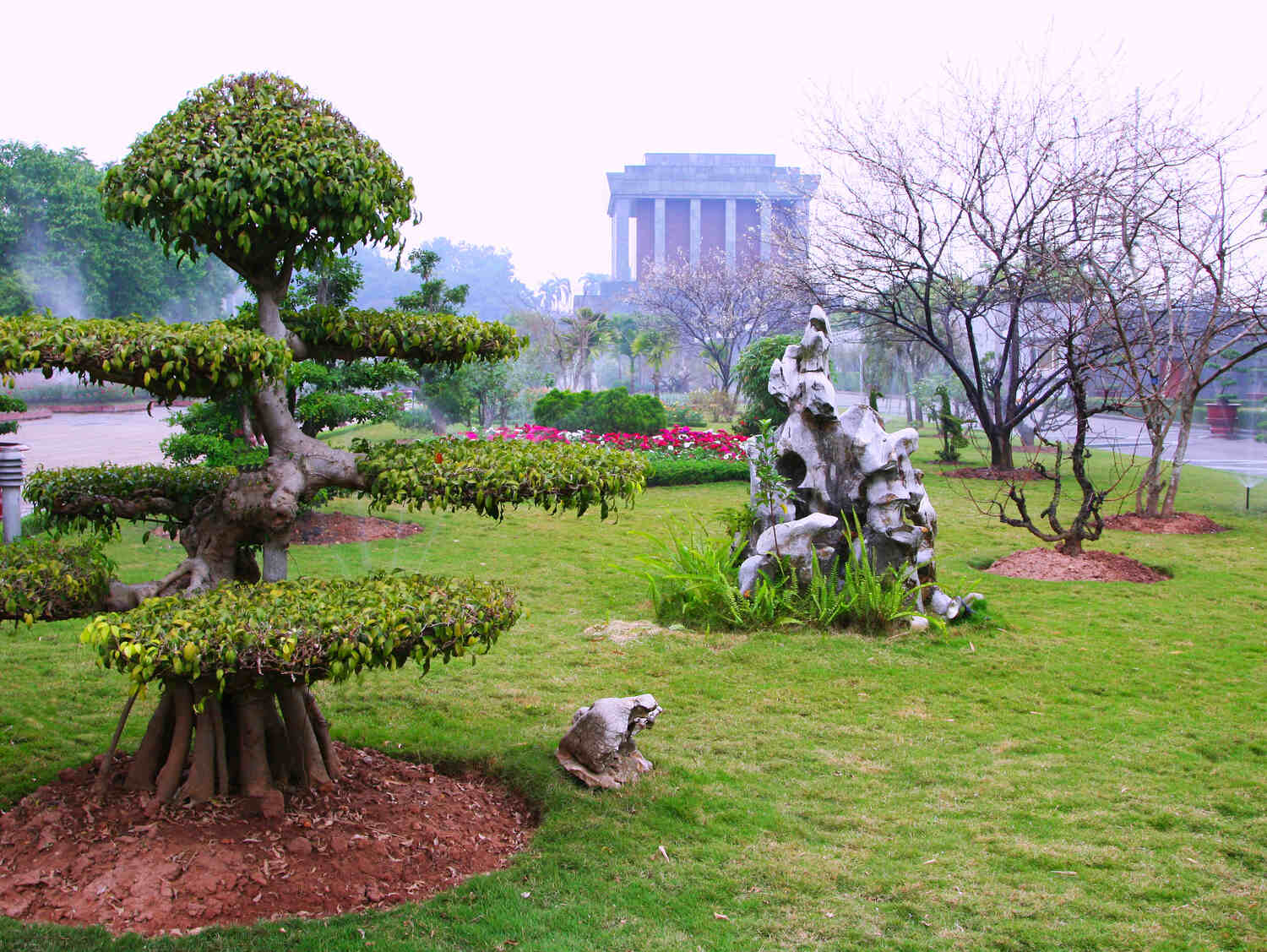 Sculpted bonsai trees and rock garden elements in a serene park with a misty Ho Chi Minh Mausoleum backdrop