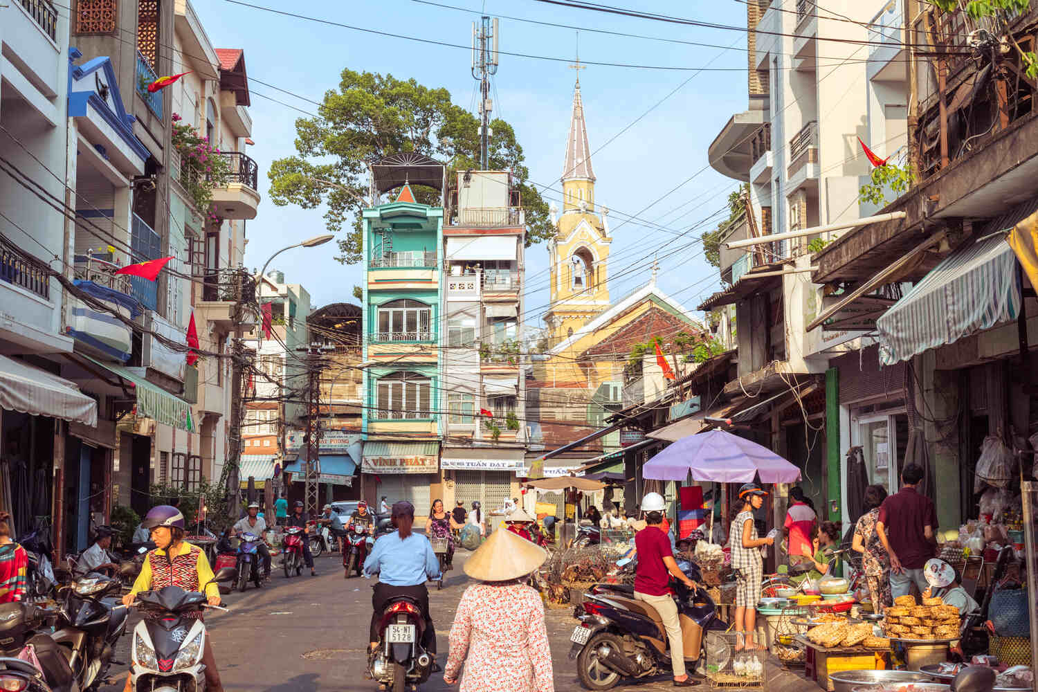 Vibrant Hanoi street scene with local businesses, pedestrians, and motorbikes under the shade of lush trees.