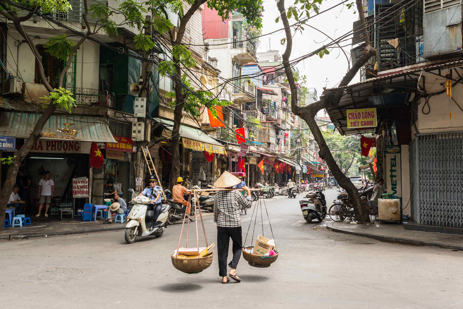 Street vendor carrying baskets on a shoulder pole, walking through a busy Hanoi street with shops and traffic.