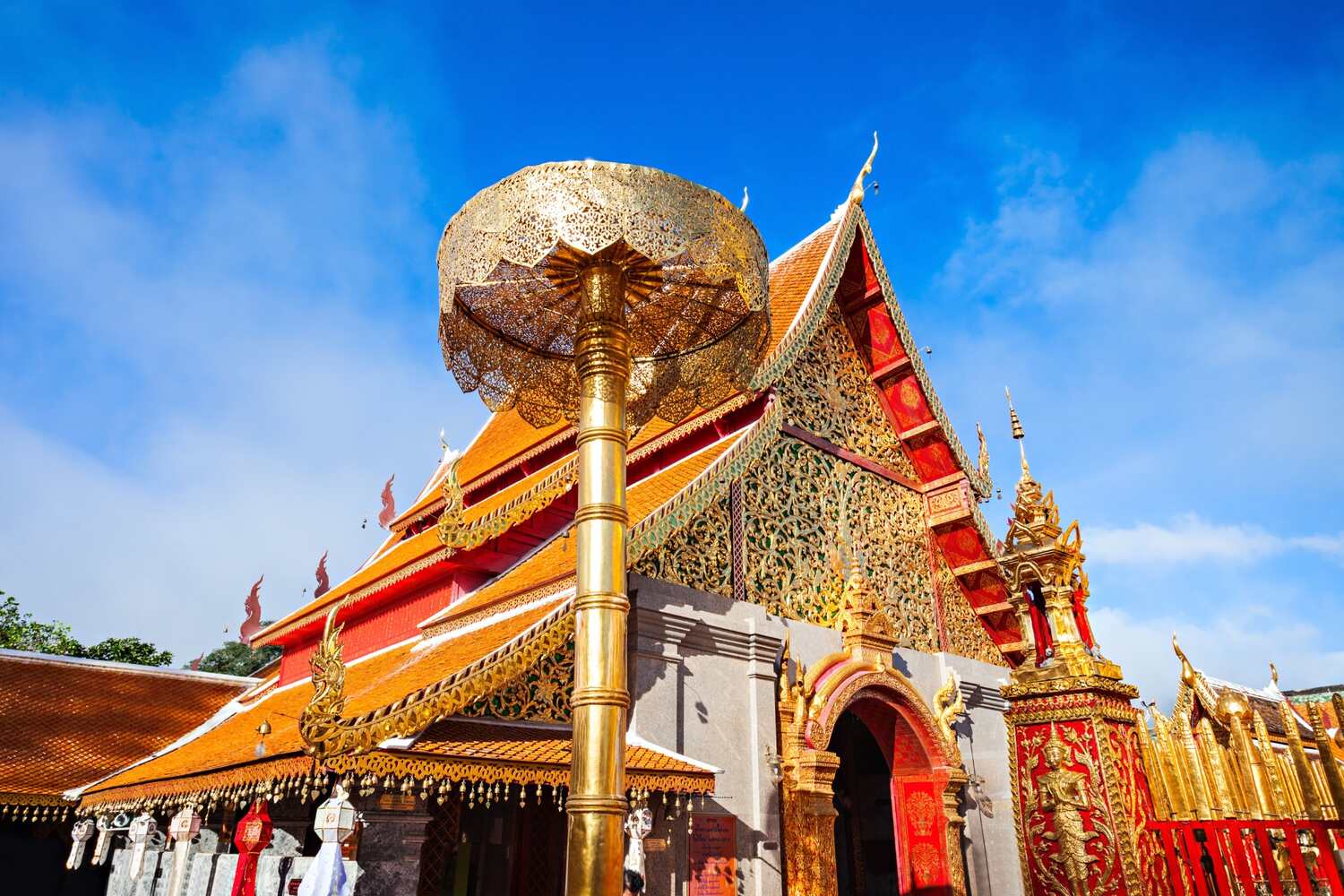 Doi suthep tours - A golden spire of a temple with intricate red and blue decorations against a blue sky with fluffy clouds.