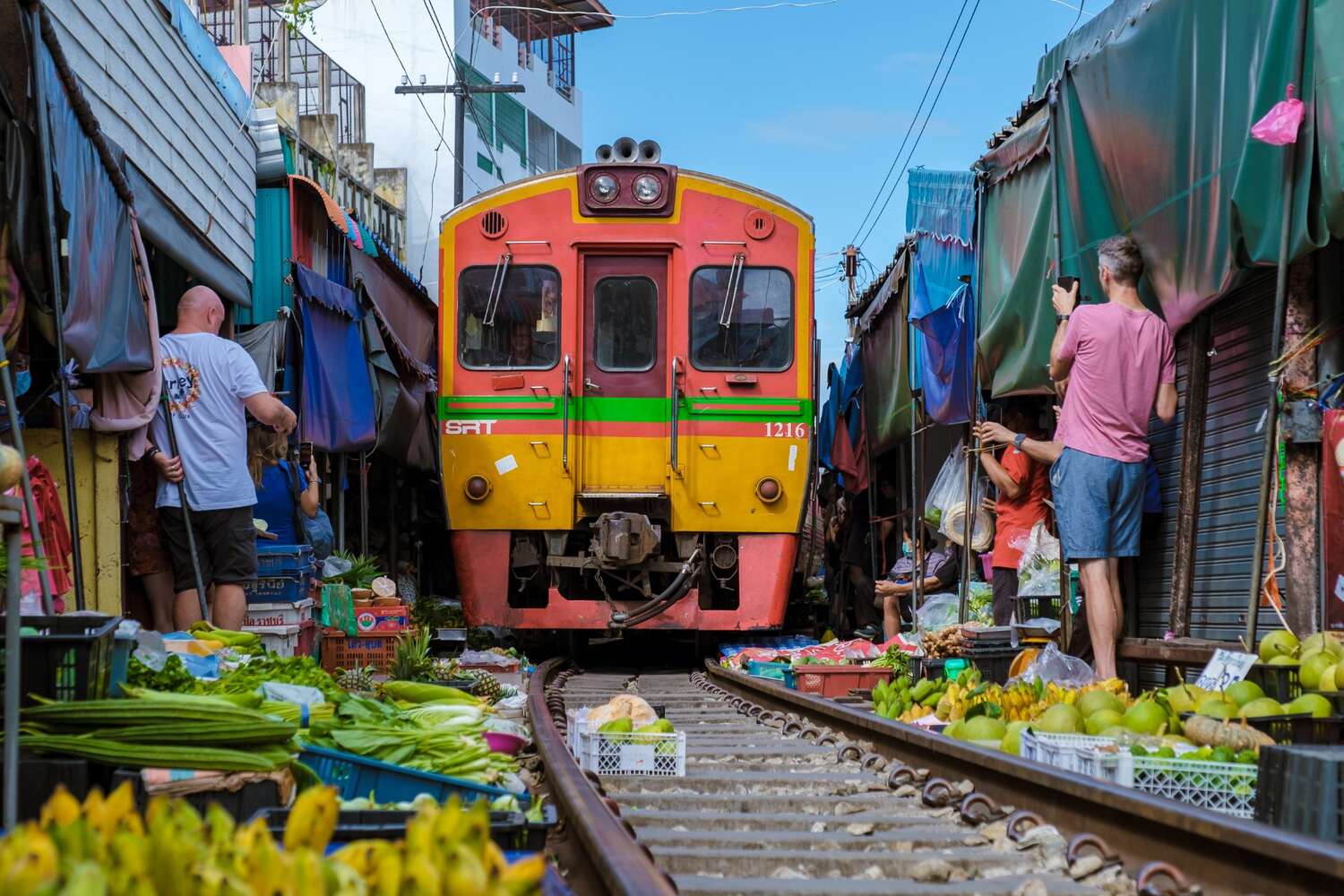 A colorful train passes through a market, with vendors and goods very close to the train tracks.