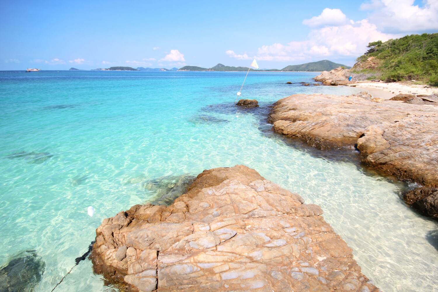 A tropical beach with clear turquoise water, rocks, and a view of distant islands under a blue sky.