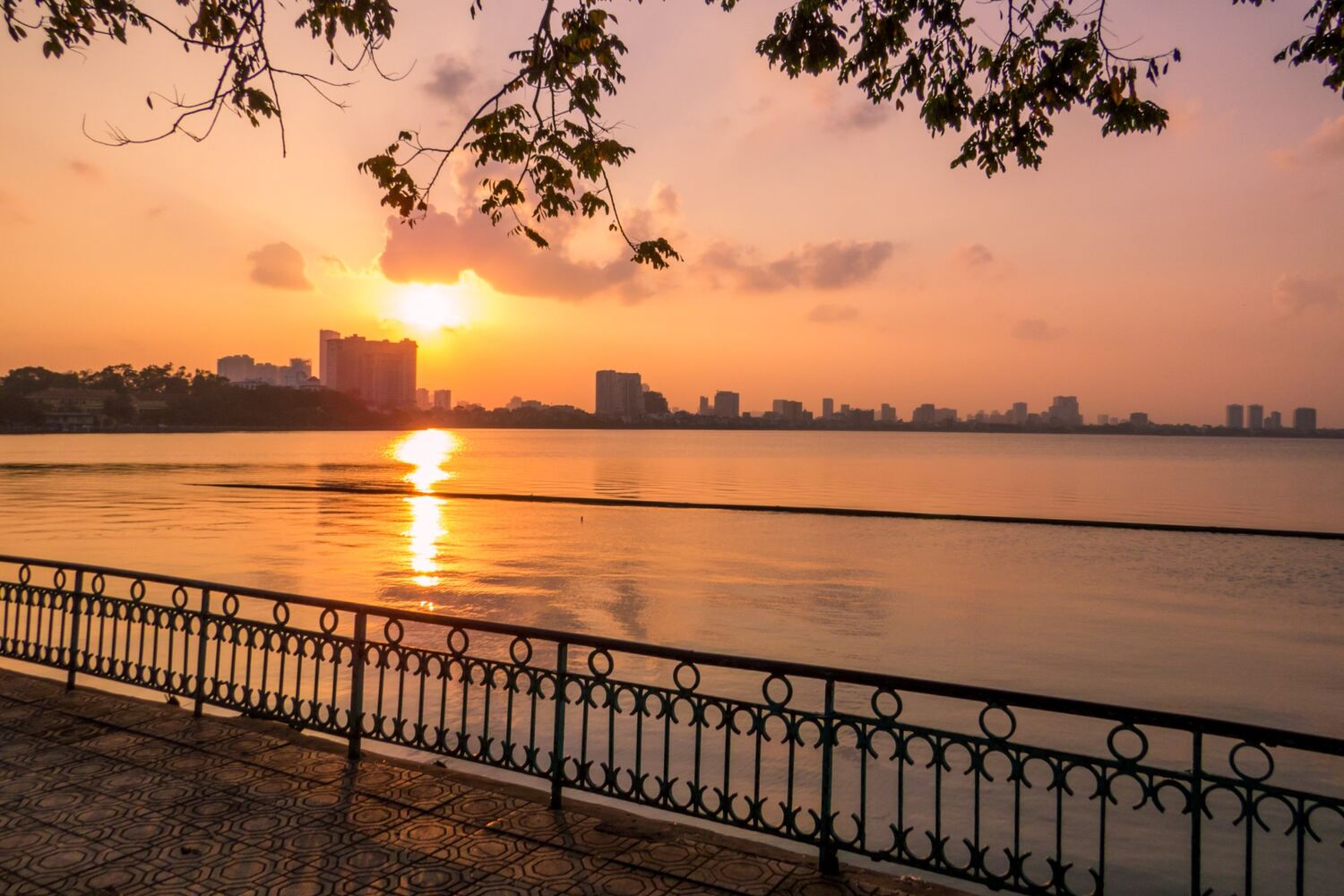 Sunset over West Lake in Hanoi, with silhouetted foliage framing the golden waters