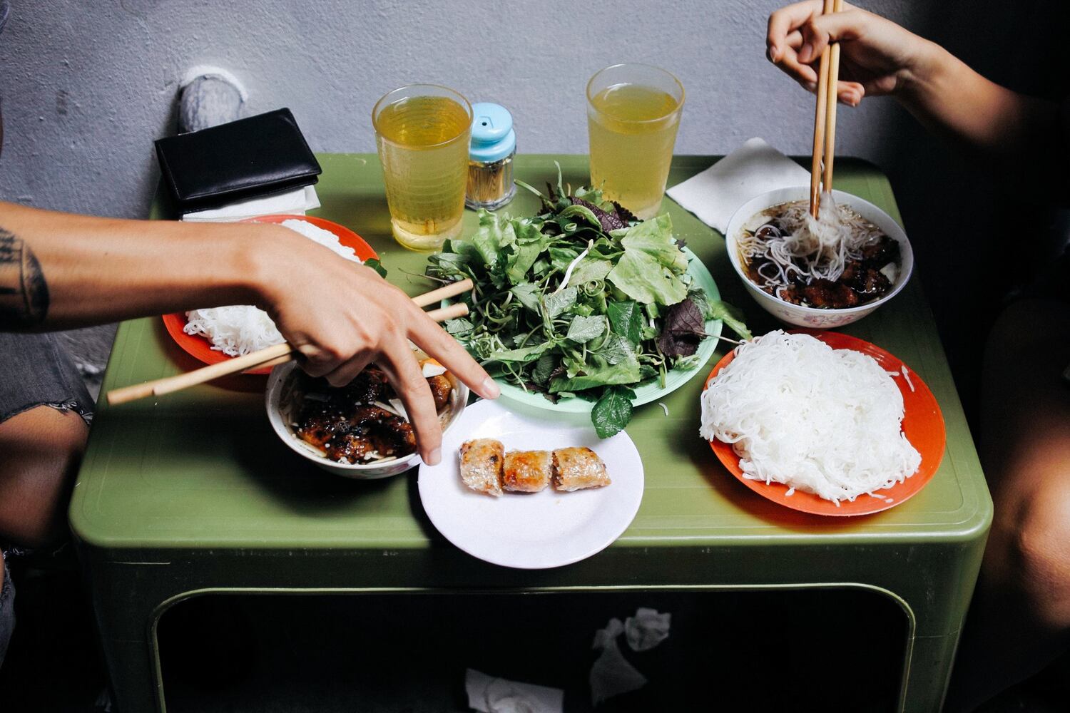 Vietnamese meal of grilled meat, fresh herbs, rice noodles served on plastic plates on a small table.