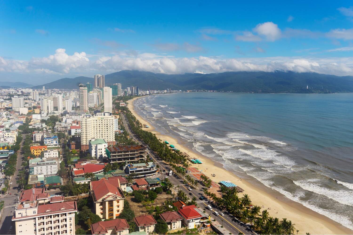 The Best 5-Day Da Nang Itinerary Travel Guide, An aerial view of a coastal city with tall buildings along a beachfront, waves approaching the shore, and a mountain range in the background.