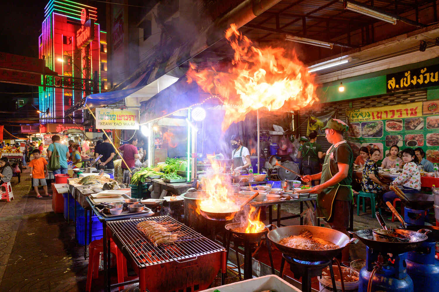 A vibrant food stall with a large flame coming from a wok, cooked by a vendor at a night market.