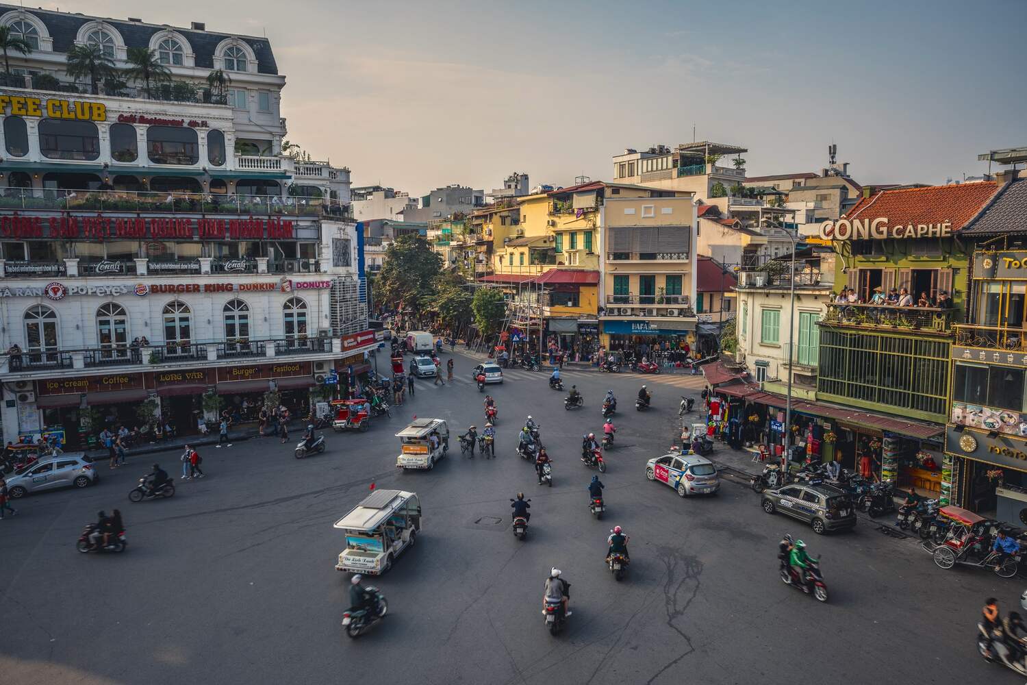 Bustling roundabout in Hanoi, Vietnam, with motorbikes and cars amid colonial and modern buildings