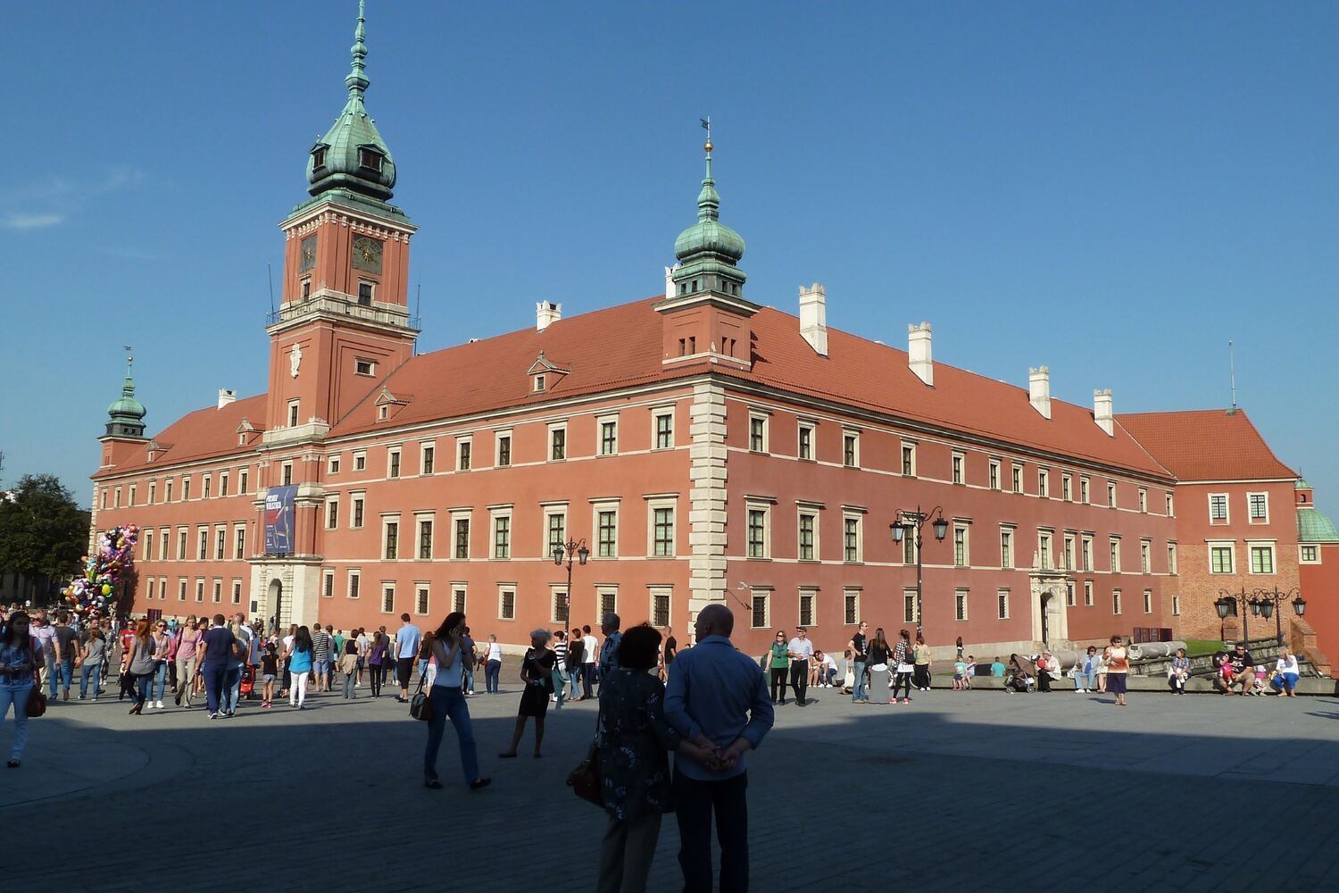 Enter the Royal Castle in Warsaw - A historic red-brick castle complex with a tower under a sunny sky.